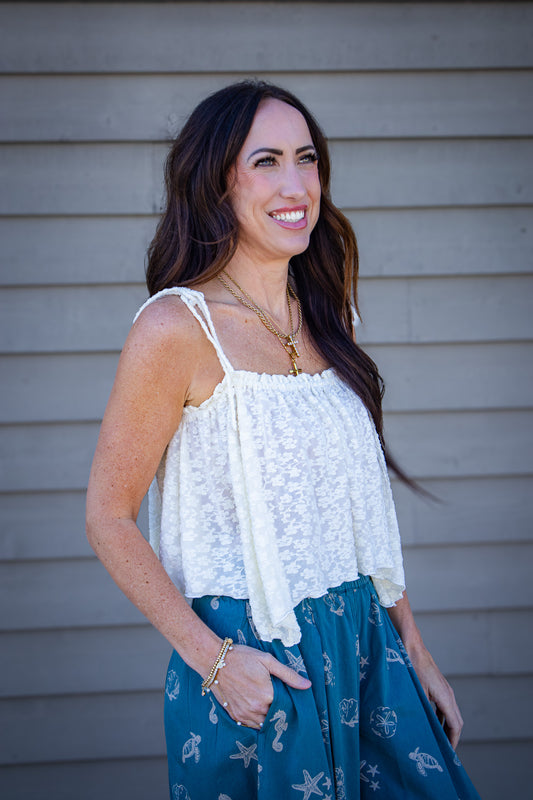 Woman wearing a cream textured tie-strap tank top paired with teal bubble pants featuring seashell and ocean print, styled with layered gold necklaces for a coastal summer outfit