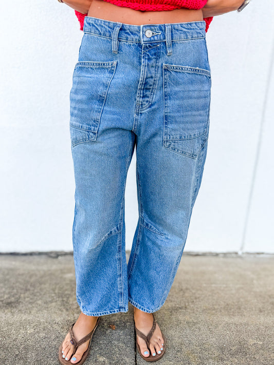 A person wearing light blue high-waisted cropped denim jeans with front patch pockets, paired with brown thong sandals and a red top, standing against a white wall.