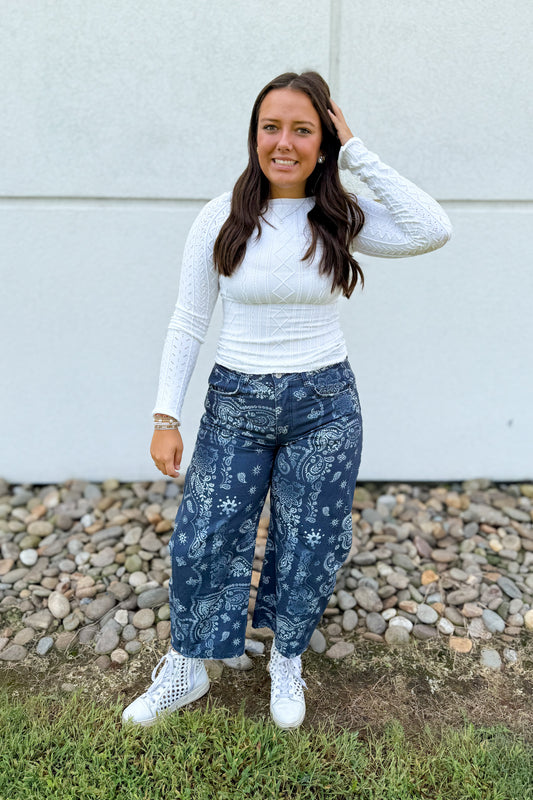 A woman stands outdoors in front of a light gray wall and gravel, wearing a white textured long-sleeve top, dark blue wide-leg jeans with a white paisley and floral pattern, and white sneakers.