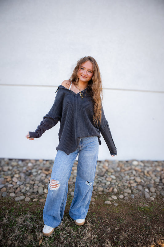 Woman wearing a dark gray oversized top and light blue, ripped jeans standing against a white wall.