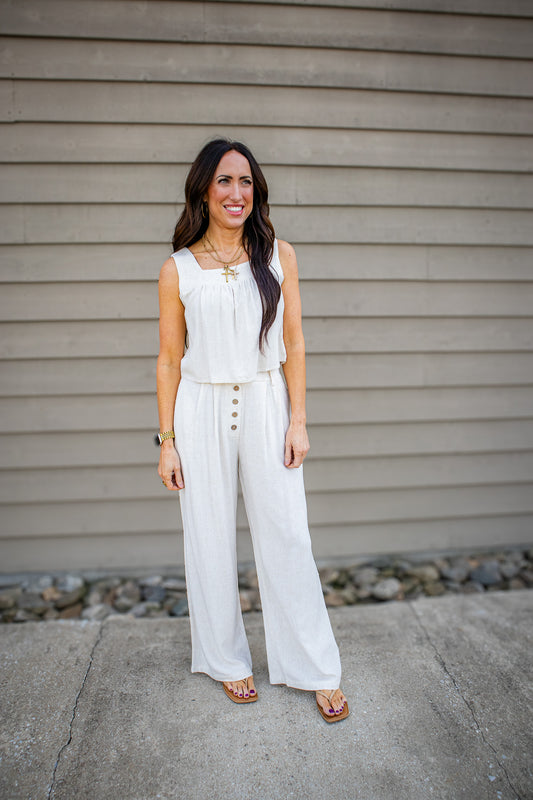 Woman wearing a white sleeveless top and wide-leg pants standing against a gray wall.
