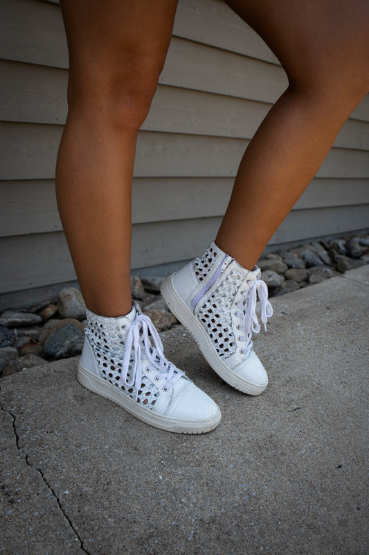 White high-top sneakers with perforated design worn by a person on a concrete surface.