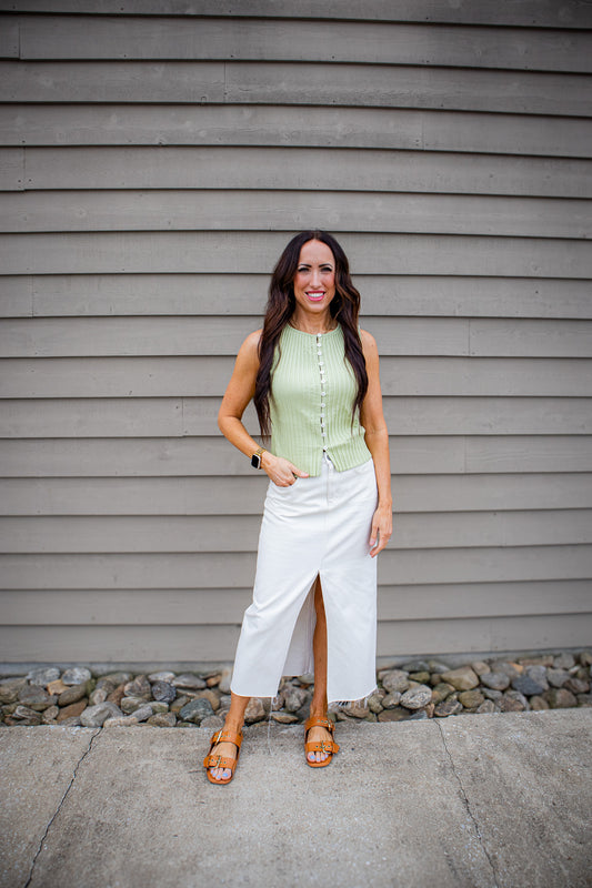 A smiling woman stands in front of a gray siding wall, wearing a light green ribbed sleeveless top with buttons and a cream midi skirt with a front slit, paired with brown sandals.