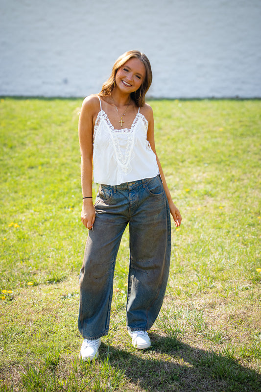 Woman wearing a white lace top and worn blue barrel jeans standing in a grassy field.