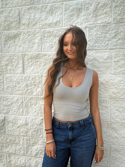 Woman wearing a gray tank top and blue jeans standing against a light stone wall.