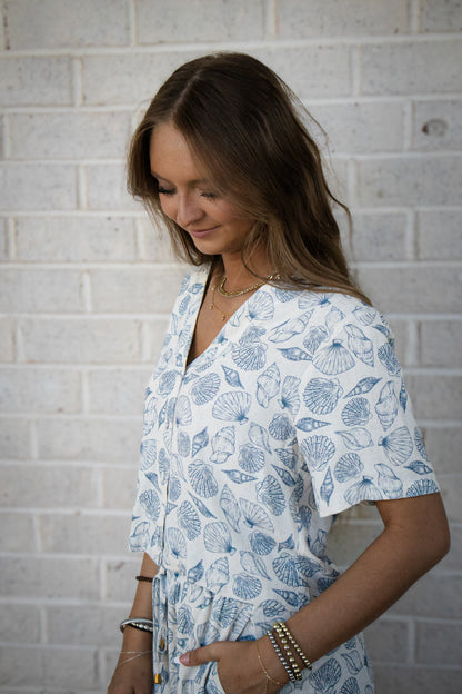 A woman wearing a seashell linen top with a v-neck, drop sleeves, and a button front detail. The top is primarily in shades of blue with a seashell pattern.