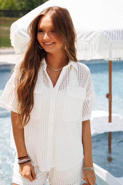 A woman standing near a pool wearing a white crochet top with a button down front, collar, and cuffed sleeves, paired with white shorts.
