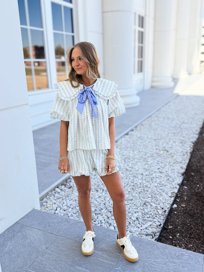 Woman wearing a light blue striped two-piece set with puff sleeves and a large blue bow detail at the neckline, styled with neutral sneakers, standing outside by white columns and gravel landscaping for a soft spring look.