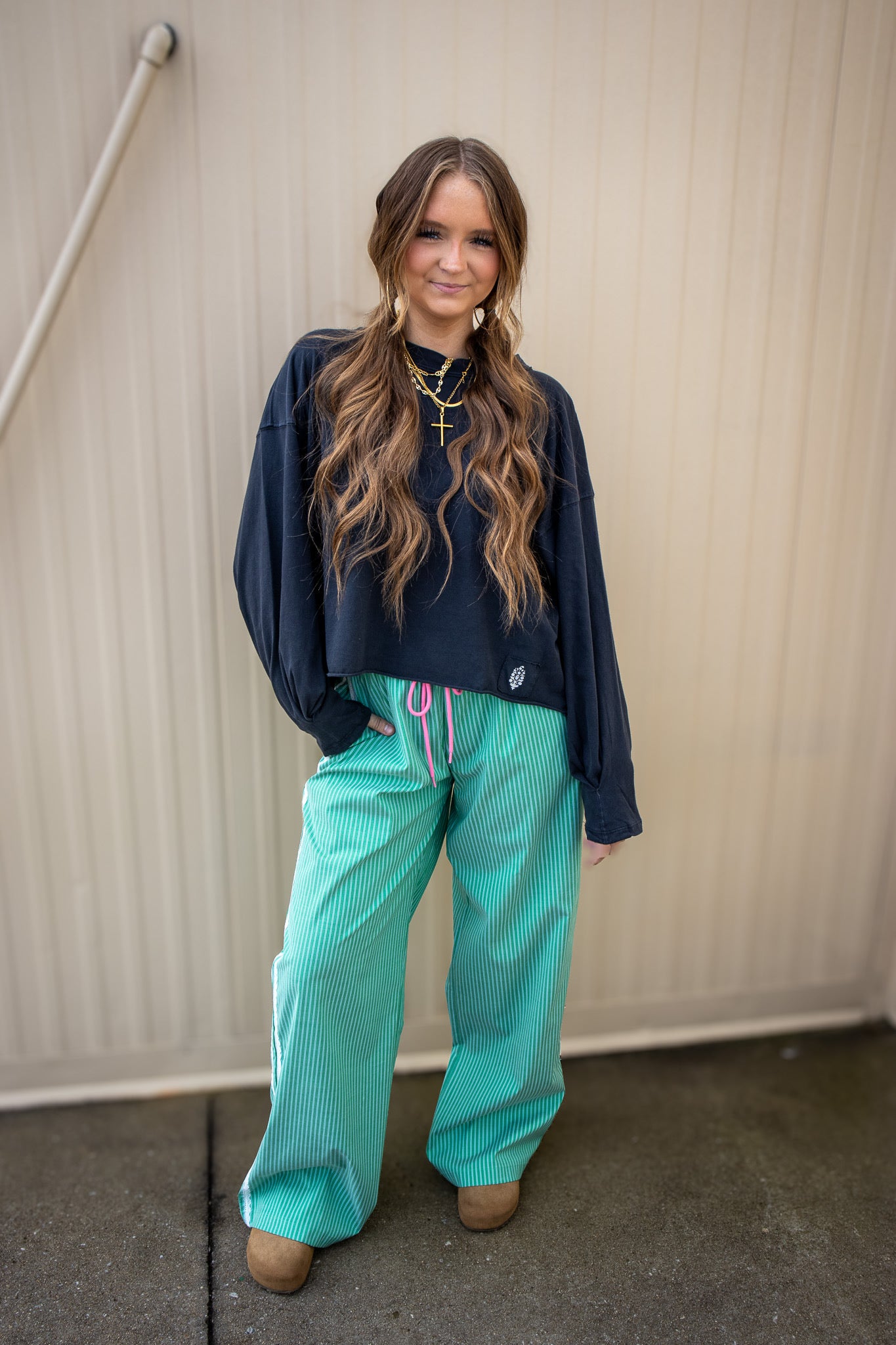 Woman wearing a navy sweater and teal pants standing against a beige wall.