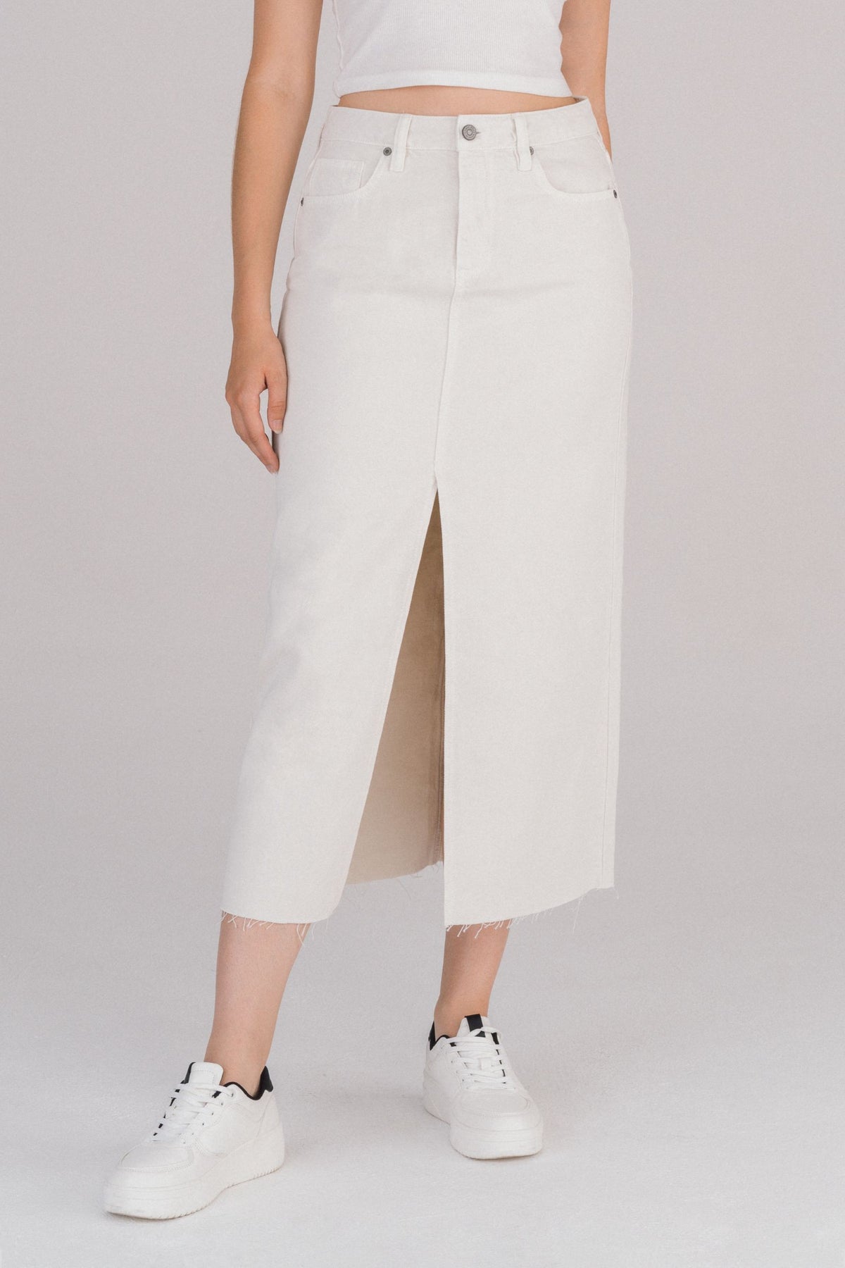 Woman wearing an off-white denim midi skirt with front slit, raw hem, and five-pocket design paired with white sneakers