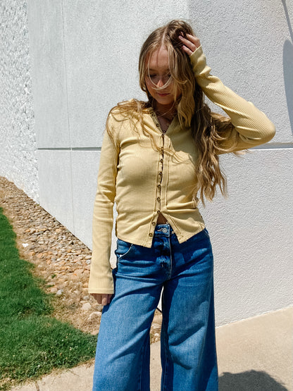 Woman wearing a yellow blouse with functional buttons and blue jeans standing against a white wall.