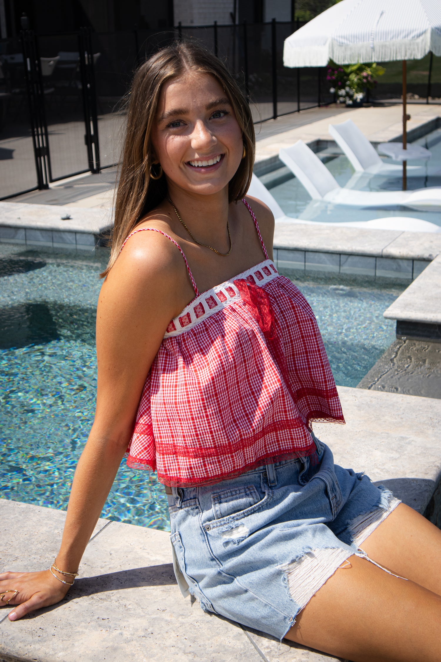 Young woman sitting by a pool wearing a red plaid top and denim shorts.