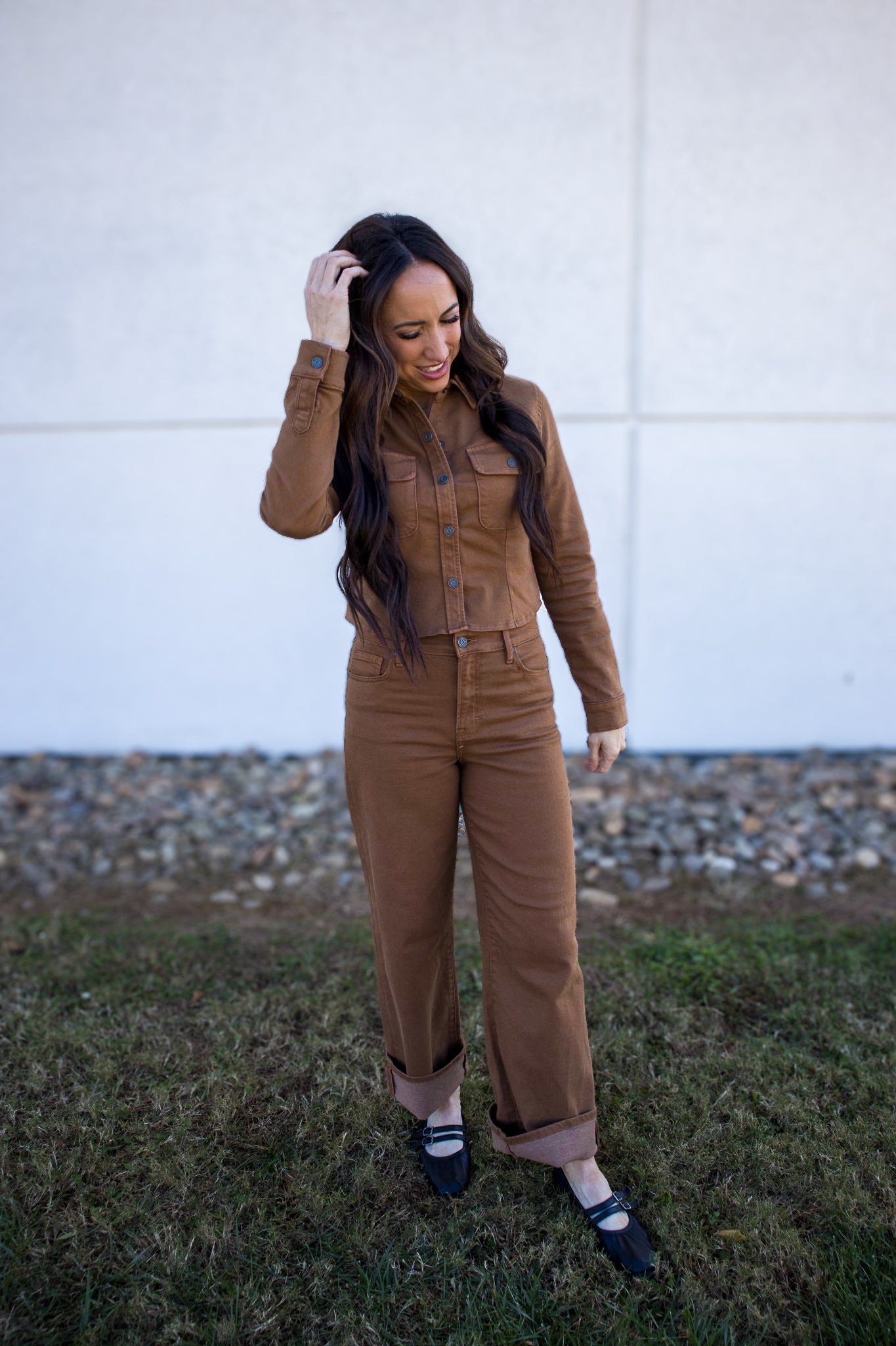 Woman wearing a brown jumpsuit standing outdoors against a white wall.