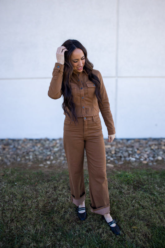 Woman wearing a brown jumpsuit standing outdoors against a white wall.