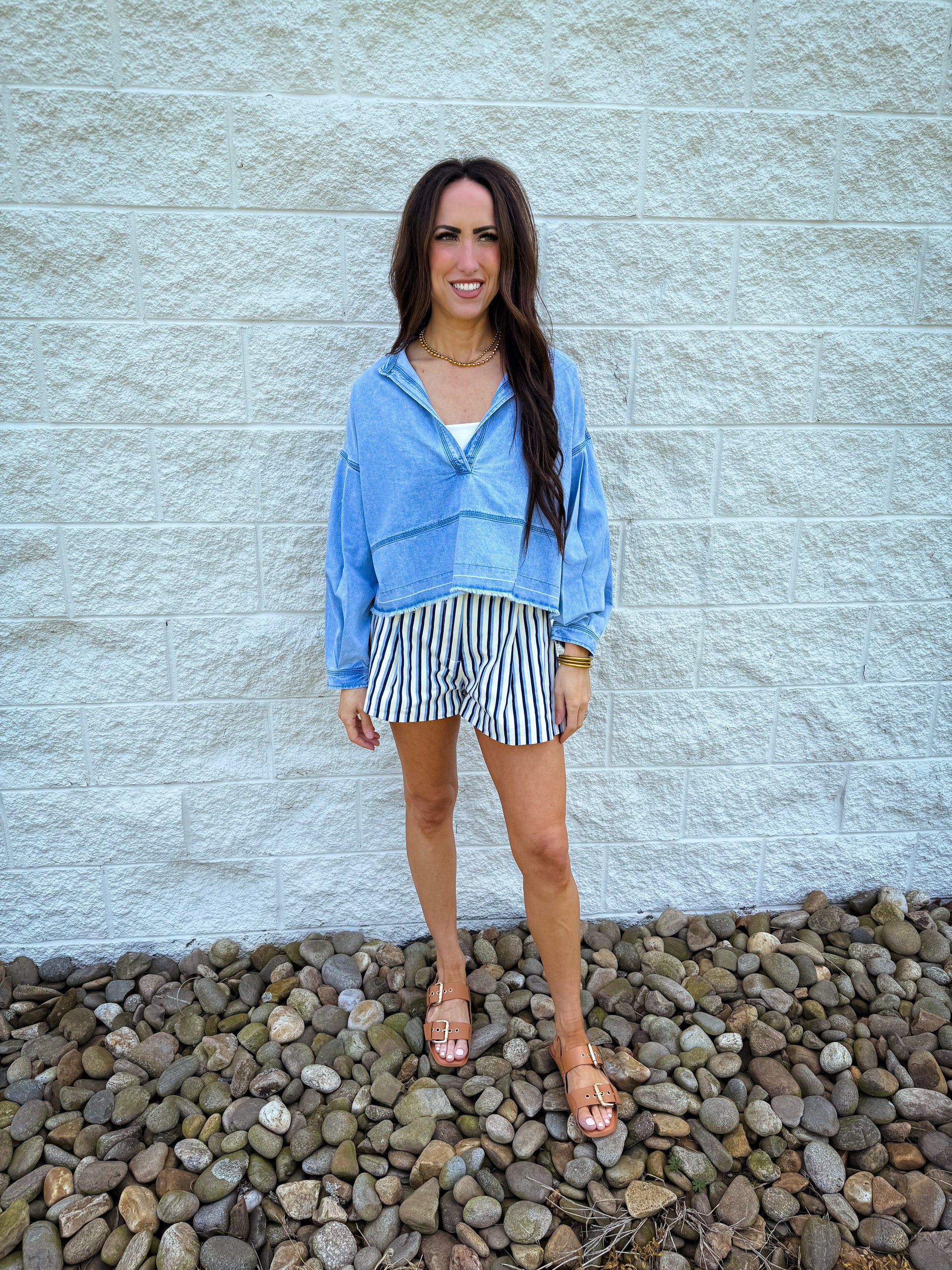 A person wearing a blue striped denim jacket and Retro Rails stripe shorts, standing in front of a white wall on a pebble surface.