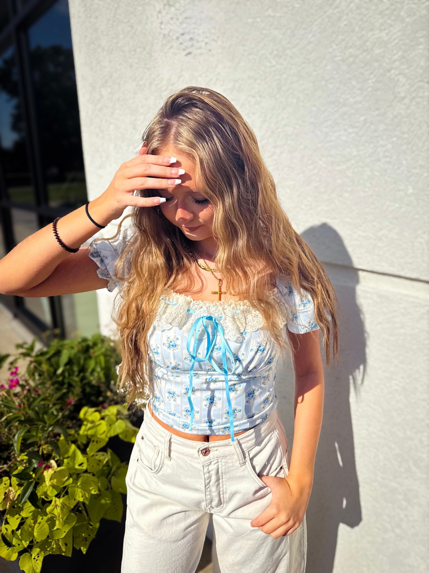 Woman wearing a light blue floral top and white pants against a gray wall.