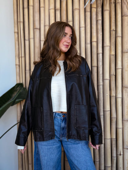 Black faux-leather zip jacket styled with a cream ribbed sweater and relaxed denim, photographed against natural bamboo for an elevated, effortless vibe.