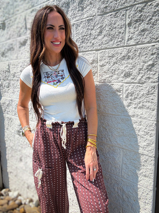 Woman wearing a white t-shirt with graphics and brown patterned pants against a stone wall.