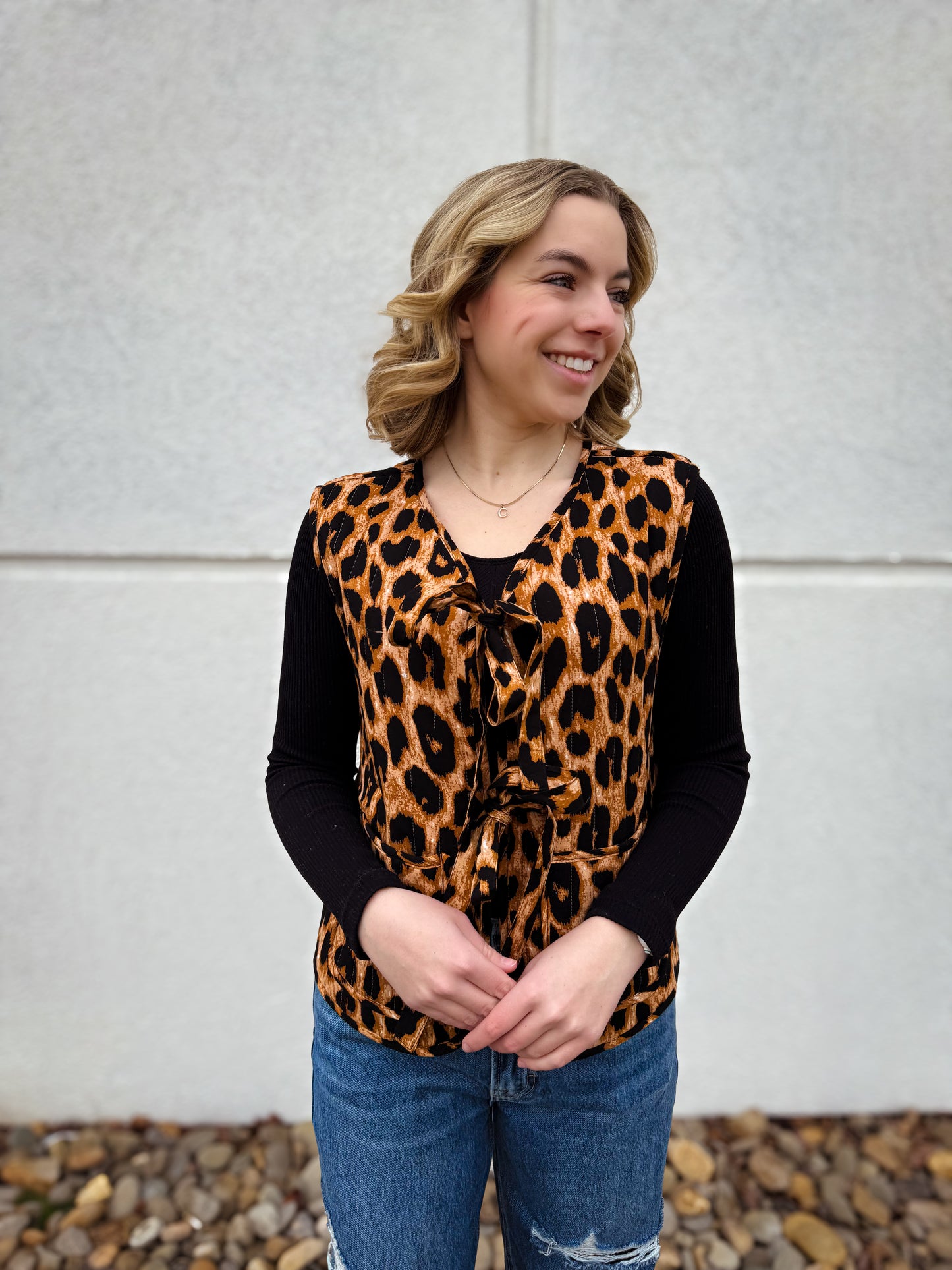 Woman wearing a leopard print vest with ties over a black long-sleeve shirt with blue jeans, standing against a light-colored wall.