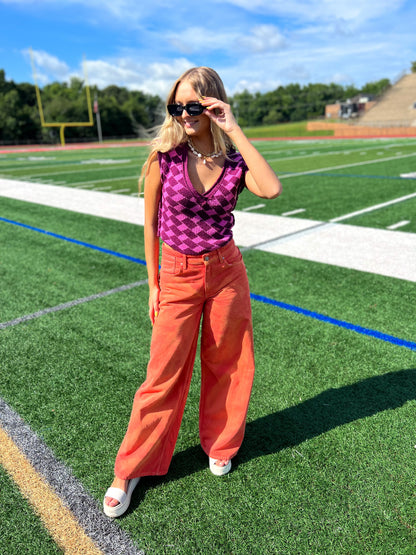 Woman in pink and purple checkered v-neck sweater and orange pants standing on a football field