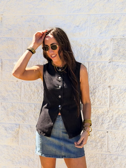 Woman wearing a black sleeveless top and denim skirt against a light stone wall.