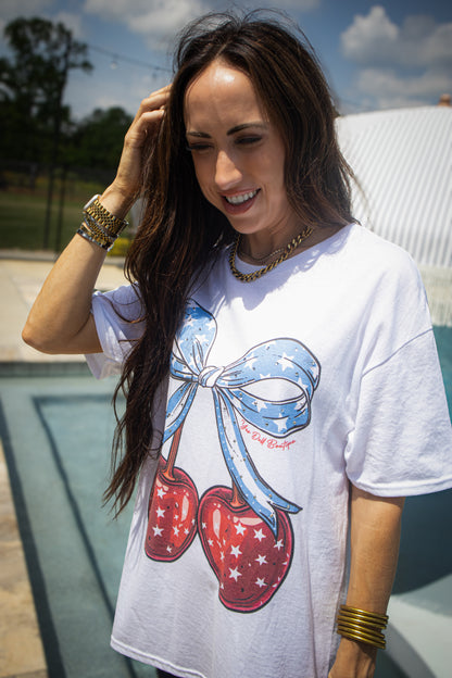 Woman smiling outdoors by a pool wearing a white graphic T-shirt with red cherries tied in a blue star-pattern bow, layered gold jewelry, and long dark hair blowing in the sun.