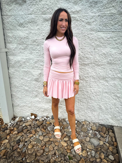 A person wearing a pink long-sleeve top and a matching pink pleated skort with a foldover waistband, standing on a rocky surface with a white wall in the background.