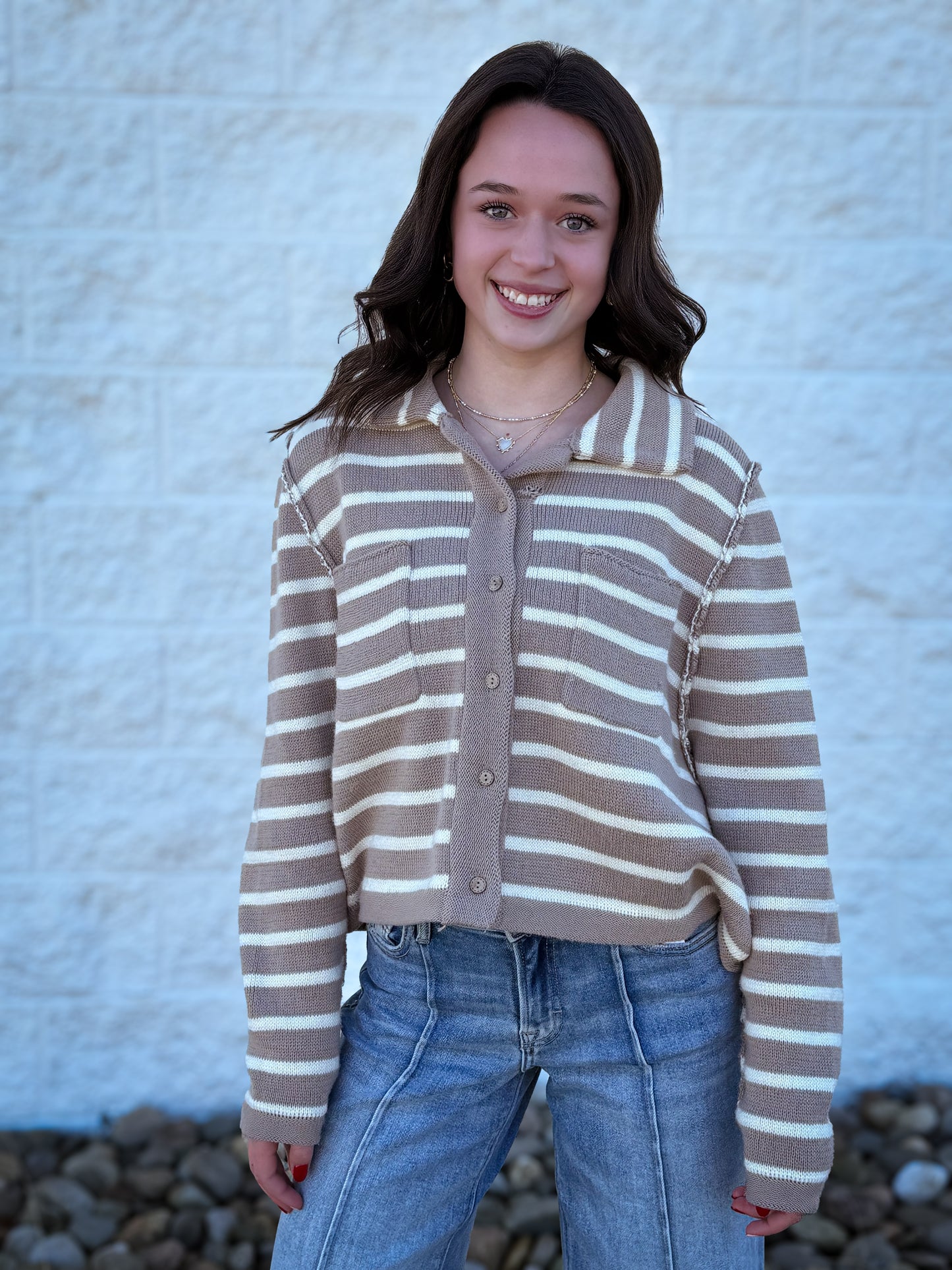 Person wearing a brown and beige striped sweater with blue jeans against a textured white wall.