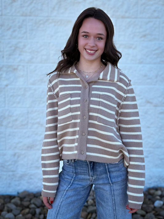 Person wearing a brown and beige striped sweater with blue jeans against a textured white wall.