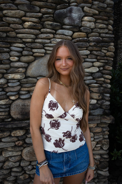 Woman wearing a white and brown floral top and denim skirt standing against a stone wall.