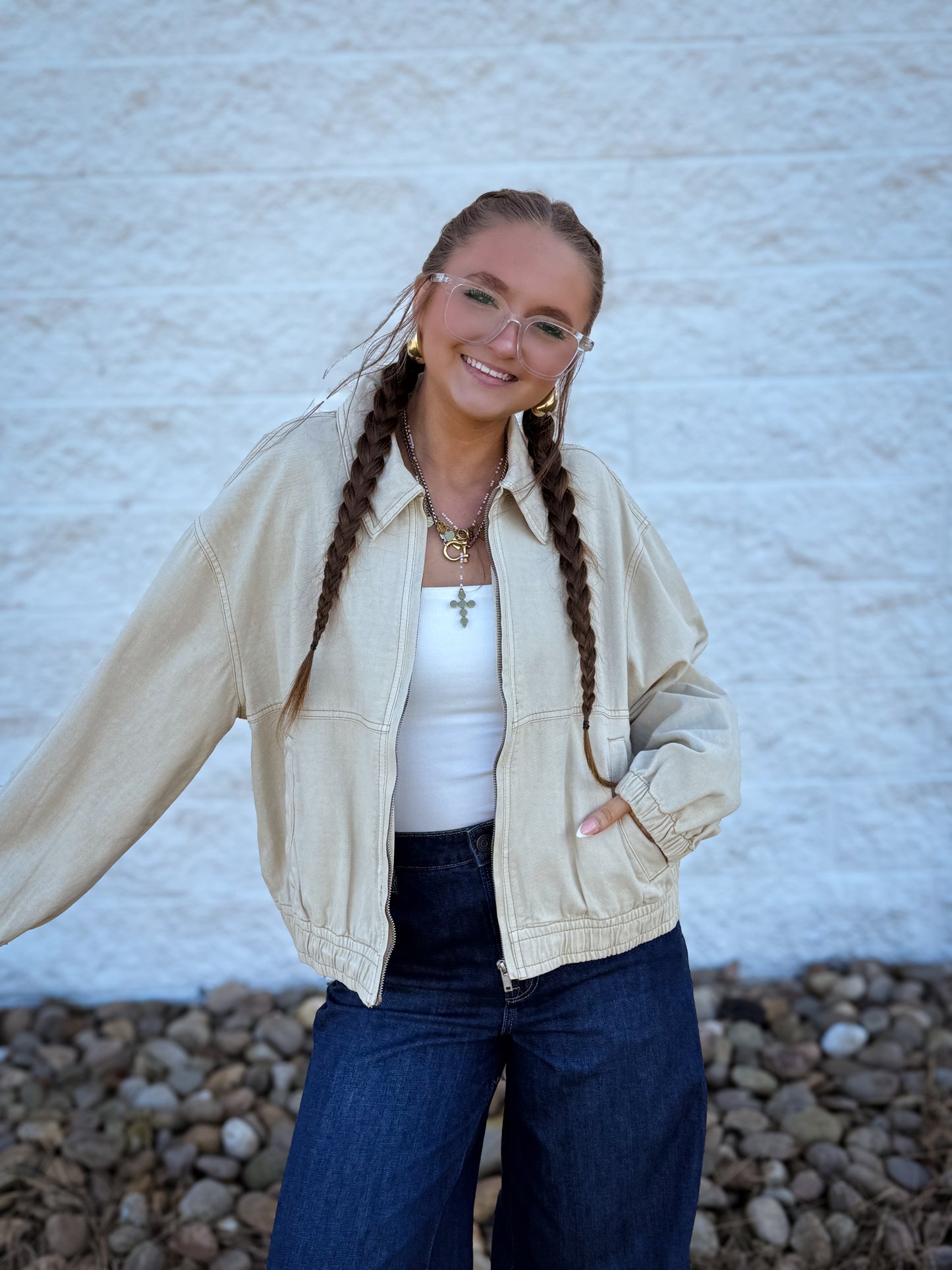 Woman with braided hair wearing a beige jacket, white top, and blue jeans against a textured wall.