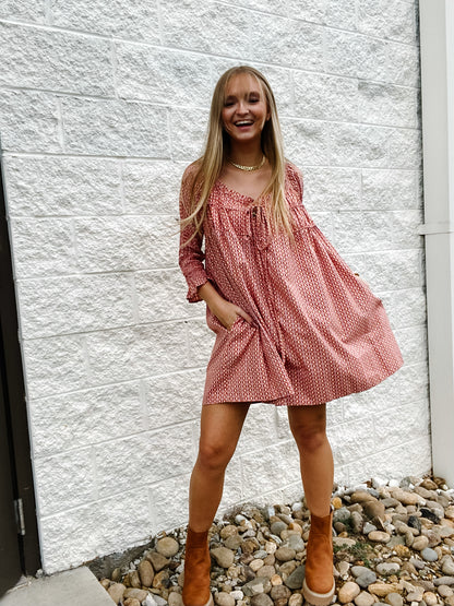 Woman in a pink, patterned dress with brown ankle boots standing against a white brick wall.