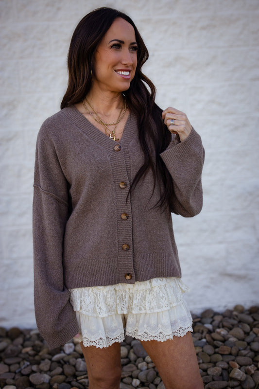 Woman wearing a brown cardigan over a white lace dress against a textured white wall.