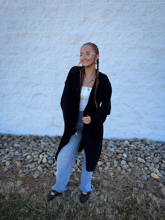 Person wearing a black coat and light blue jeans standing against a white wall.