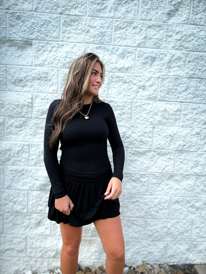 Woman wearing a black long-sleeve top and black skirt standing against a light gray brick wall.