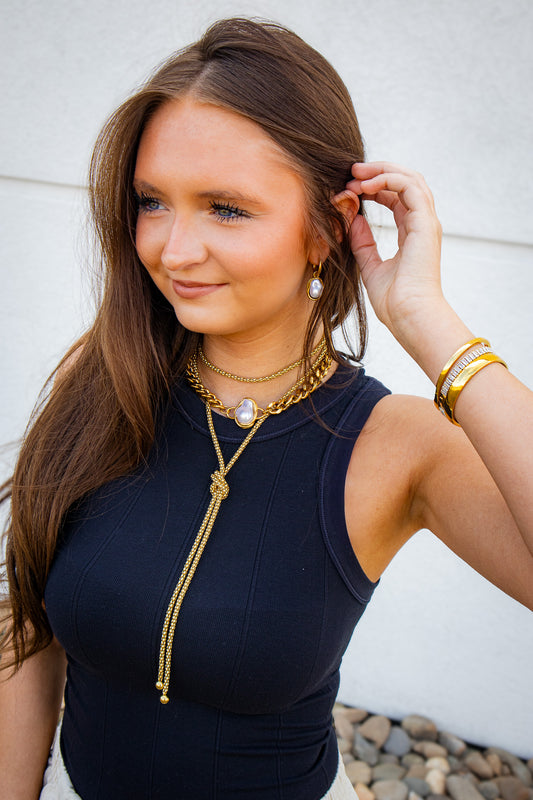 Model wearing a waterproof gold long rope knot lariat necklace featuring a textured knot detail and cascading chain tassels. Tarnish-resistant statement necklace with a lifetime warranty, layered with pearl-accented gold jewelry.