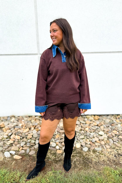A woman modeling a brown long-sleeve sweater with blue denim collar, cuffs, and hem, paired with black lace shorts and black knee-high boots, standing outdoors against a white wall and gravel background.