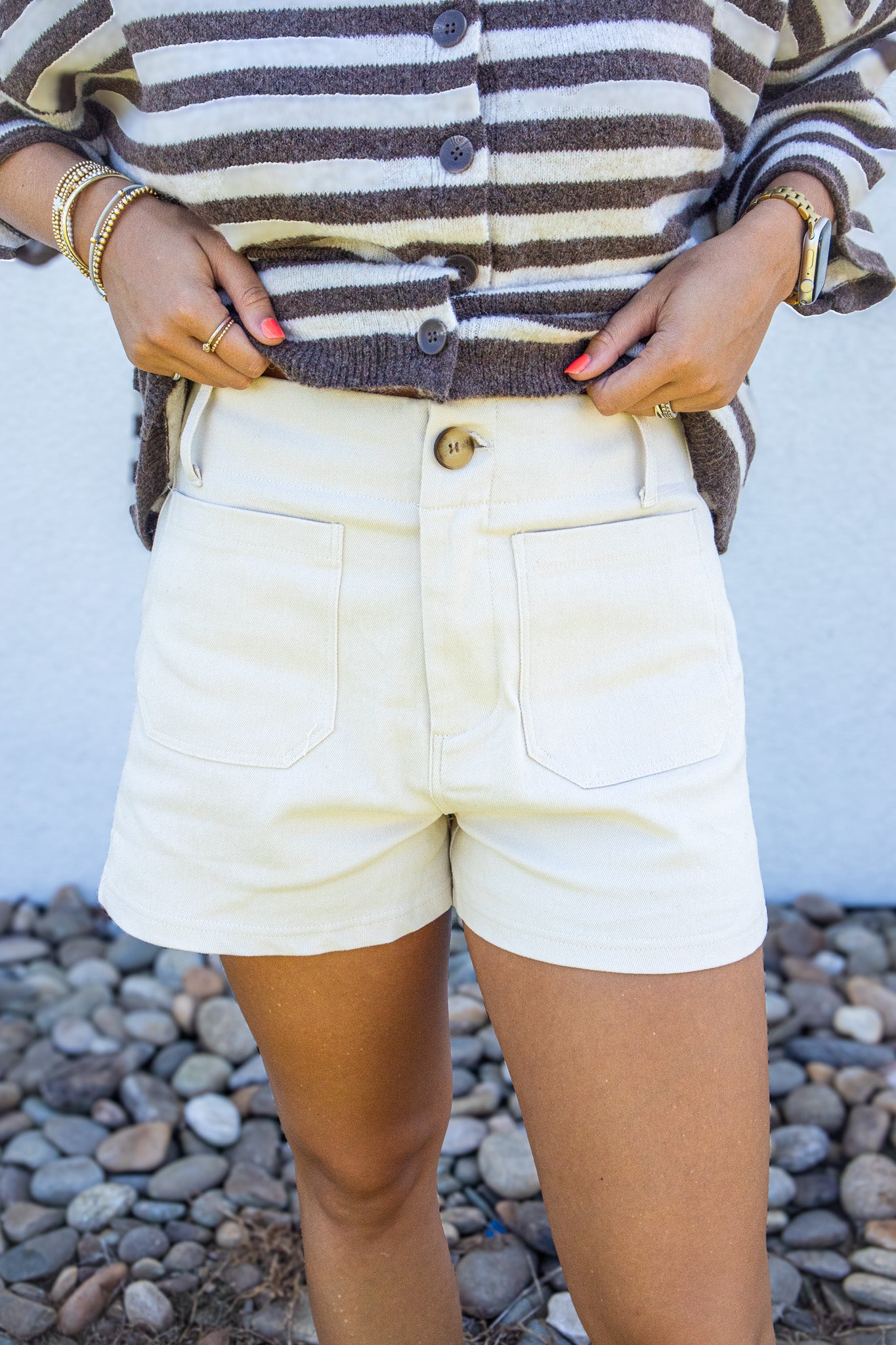 A person wearing cream-colored shorts with a brown and cream striped button-up cardigan, standing outdoors against a white wall and a bed of small gray pebbles.
