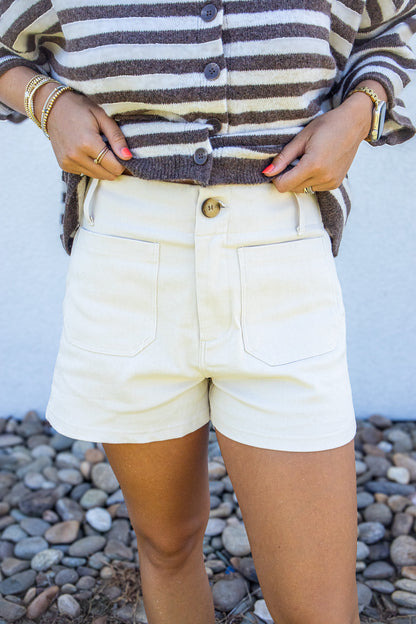 A person wearing cream-colored shorts with a brown and cream striped button-up cardigan, standing outdoors against a white wall and a bed of small gray pebbles.