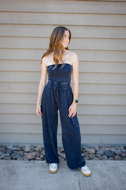 Women’s dark mineral wash strapless wide leg jumpsuit with smocked drawstring waist styled with sneakers.