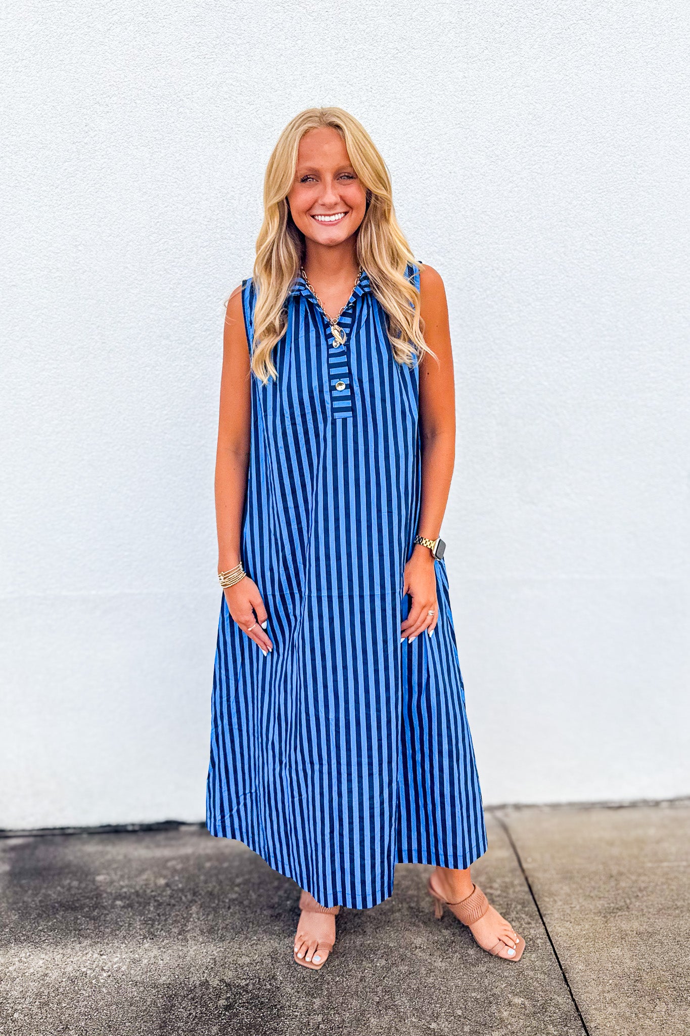 A woman modeling a sleeveless blue and white vertically striped midi dress with a buttoned collar, standing against a white stucco wall.