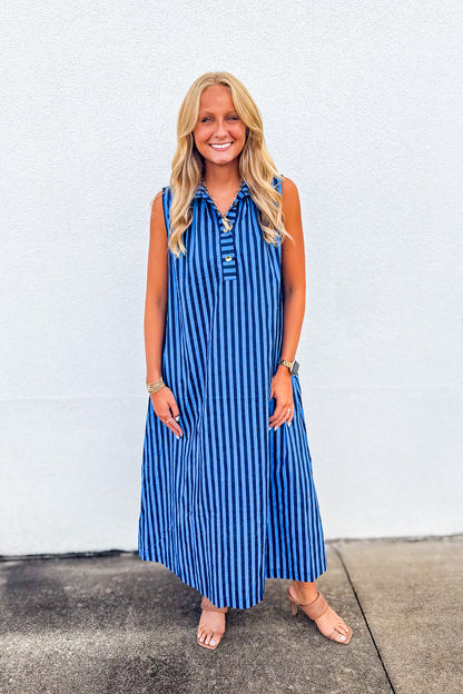 A woman modeling a sleeveless blue and white vertically striped midi dress with a buttoned collar, standing against a white stucco wall.