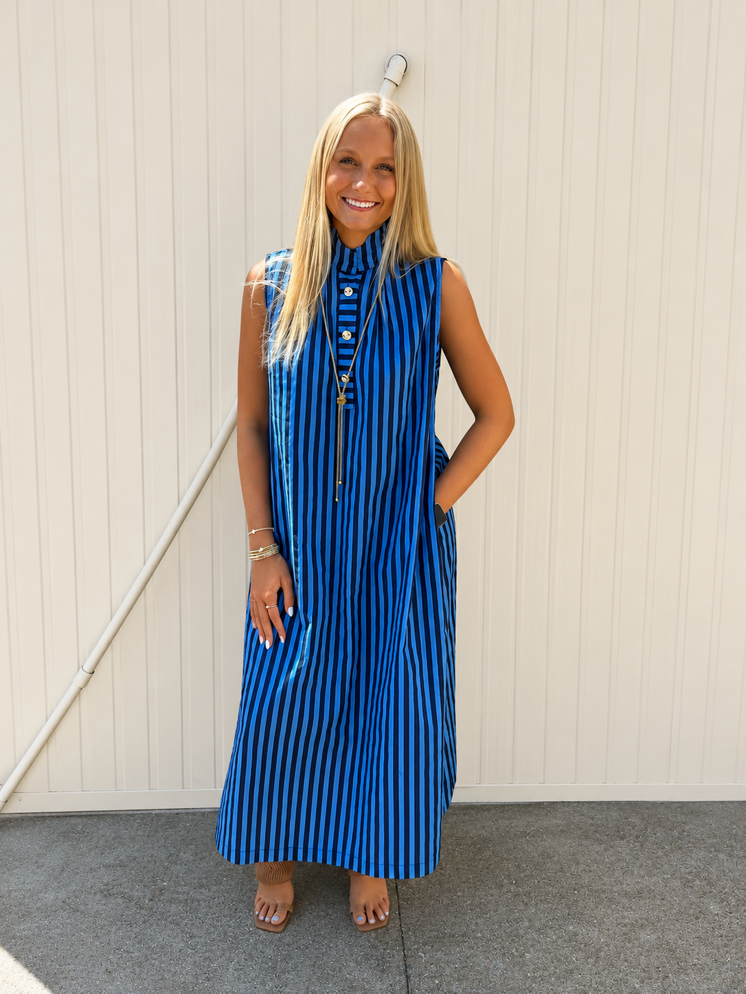 Women’s blue striped sleeveless maxi dress with a mock neckline, gold button details, side pockets, and a relaxed fit, styled with gold jewelry and neutral heels for a polished fall boutique outfit.