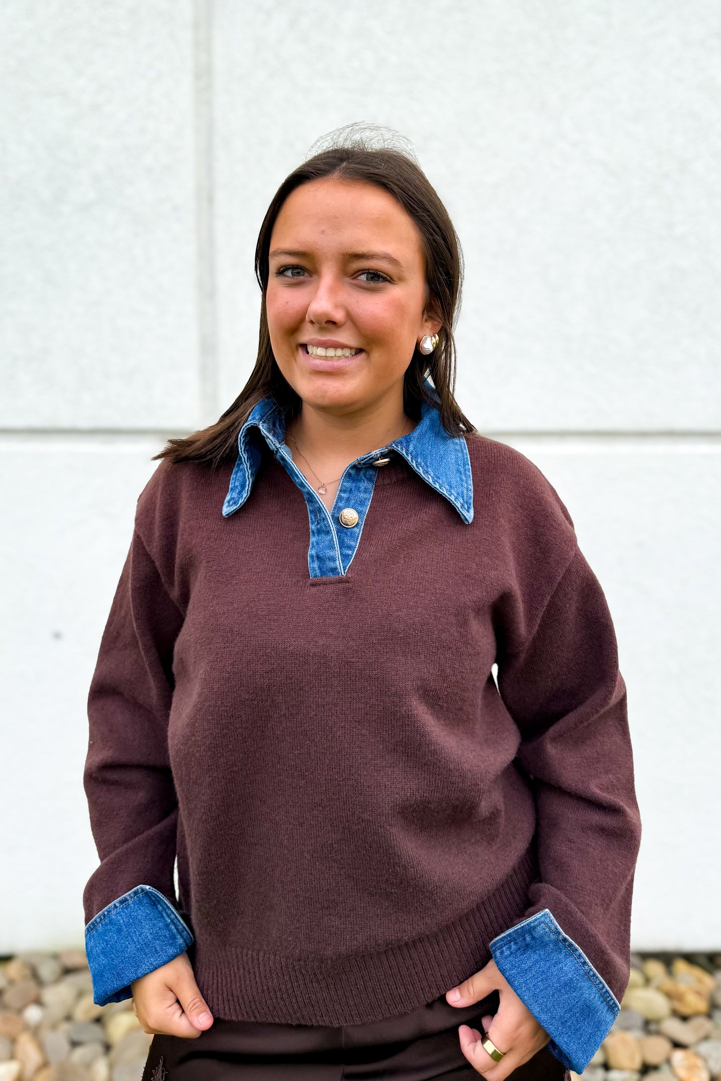A woman wearing a dark brown knit sweater with a blue denim collar and matching blue denim cuffs, standing in front of a light gray concrete wall.