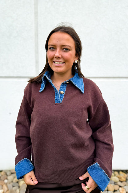 A woman wearing a dark brown knit sweater with a blue denim collar and matching blue denim cuffs, standing in front of a light gray concrete wall.