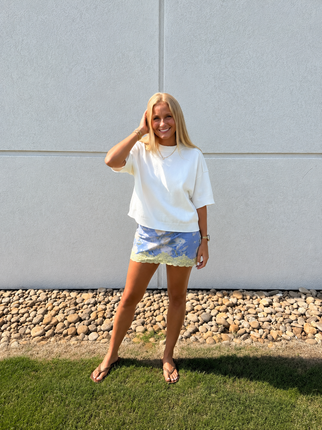Women’s white short-sleeve knit sweater paired with a blue floral lace-trim mini skirt, styled with flip-flops and gold jewelry for a chic summer-to-fall transitional boutique outfit.