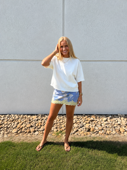 Women’s white short-sleeve knit sweater paired with a blue floral lace-trim mini skirt, styled with flip-flops and gold jewelry for a chic summer-to-fall transitional boutique outfit.
