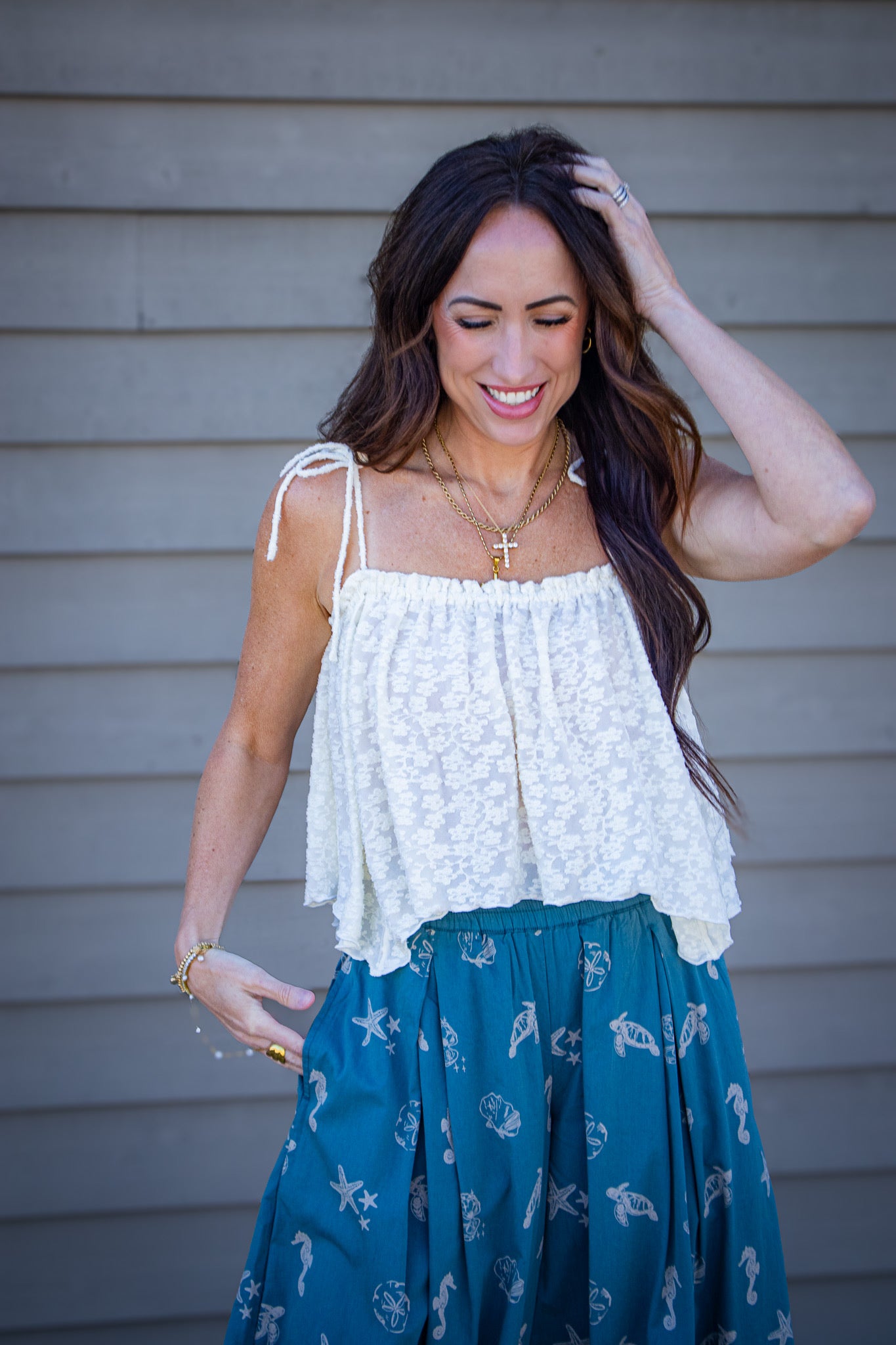 Woman wearing a cream textured tie-strap tank top paired with teal bubble pants featuring seashell and ocean print, styled with layered gold necklaces for a coastal summer outfit
