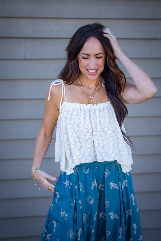 Woman wearing a cream textured tie-strap tank top paired with teal bubble pants featuring seashell and ocean print, styled with layered gold necklaces for a coastal summer outfit