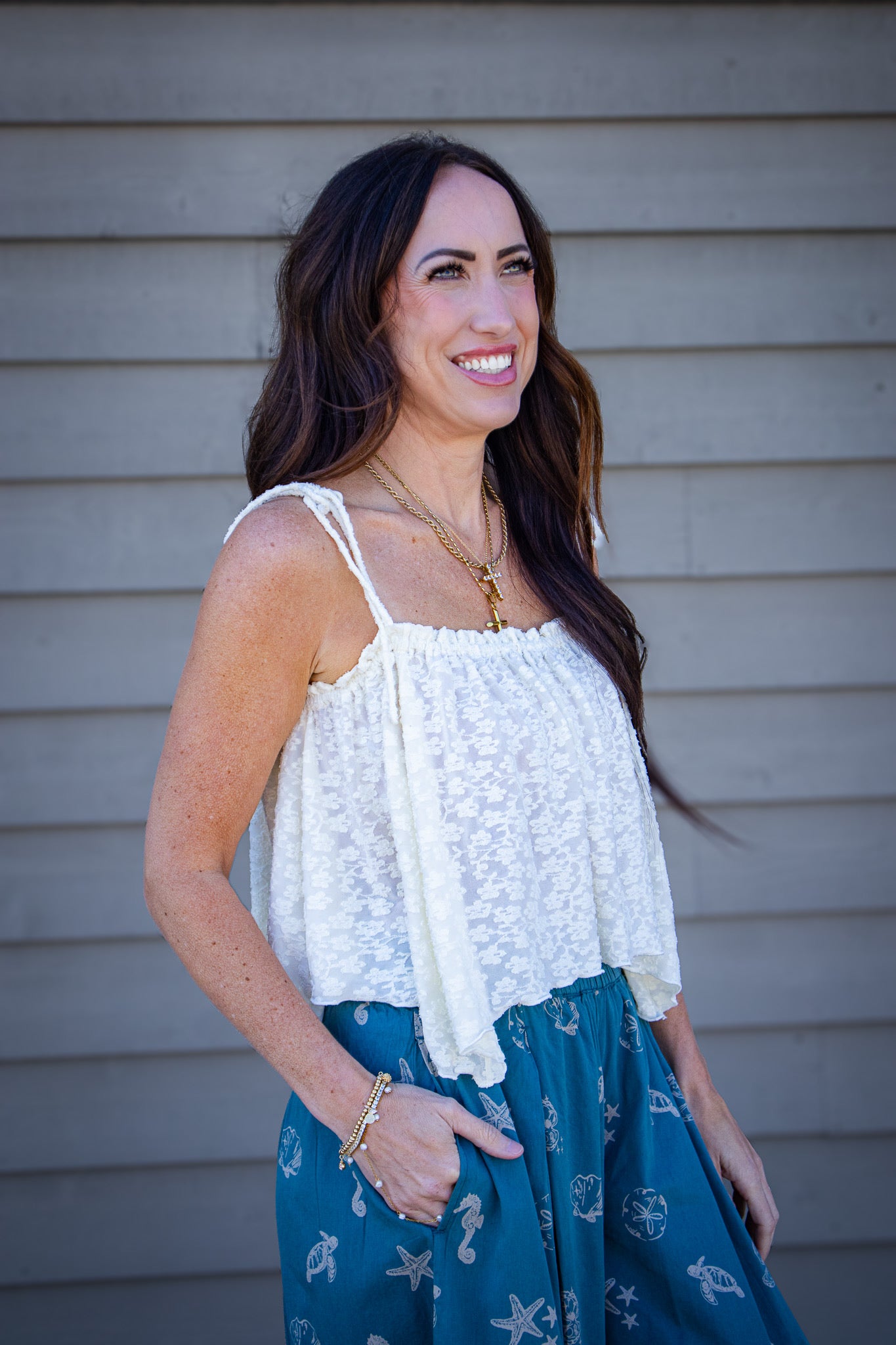 Woman wearing a cream textured tie-strap tank top paired with teal  bubble pants featuring seashell and ocean print, styled with layered gold necklaces for a coastal summer outfit